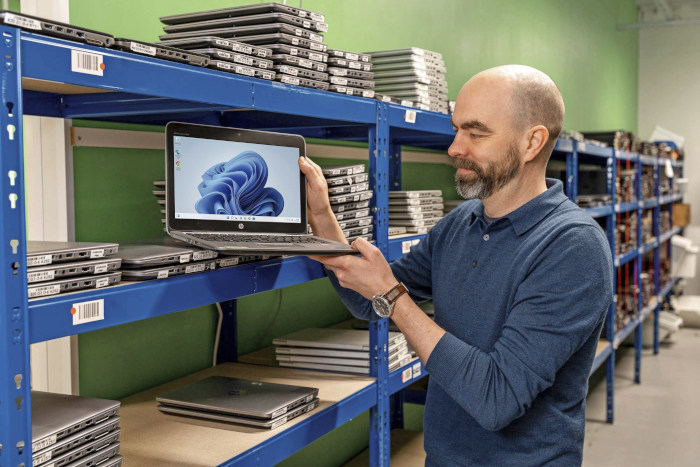 Mikko Veijola esittelee tietokonetta varastolla, jossa hyllyissä paljon samanlaisia, kierrätettyjä koneita.